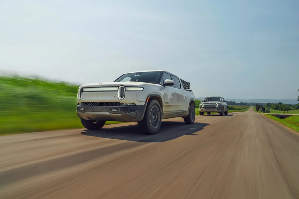 Two silver Rivian electric pick-up trucks driving down road with blurry grass on roadside